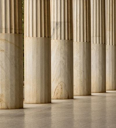 A row of classical fluted stone columns with smooth bases, lit by natural light casting shadows on the floor.
