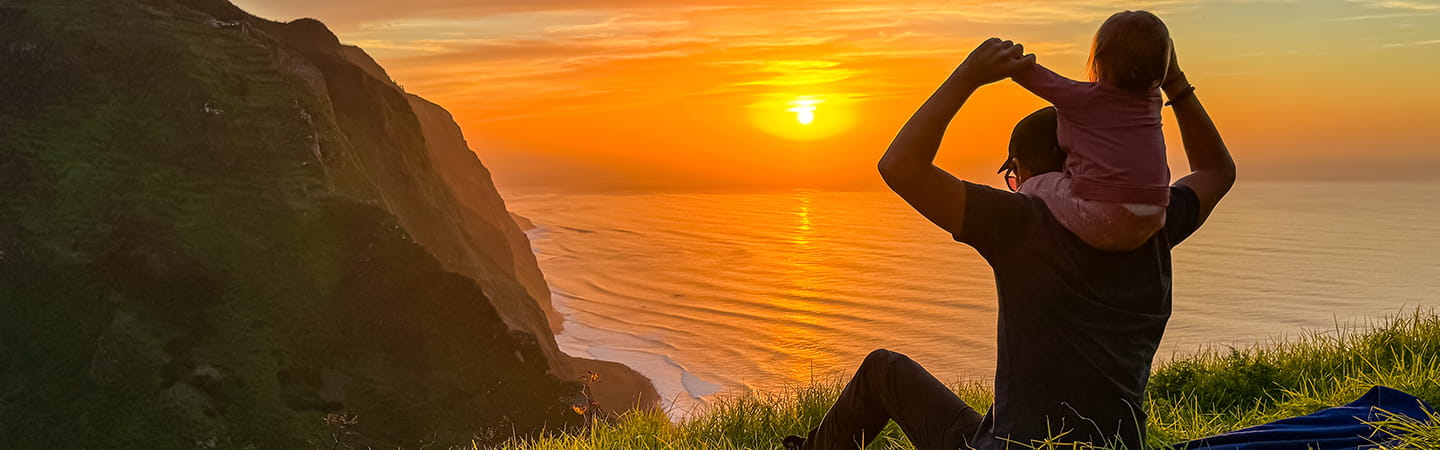 A man and his child sit on a grassy hill, gazing at the ocean under a clear blue sky.