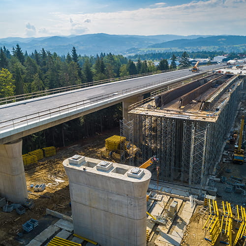 Aerial view of a highway bridge under construction over a forested area with mountains visible in the background.