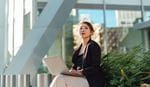 A woman in business attire sits outdoors with a laptop, surrounded by plants, beneath modern architecture. 