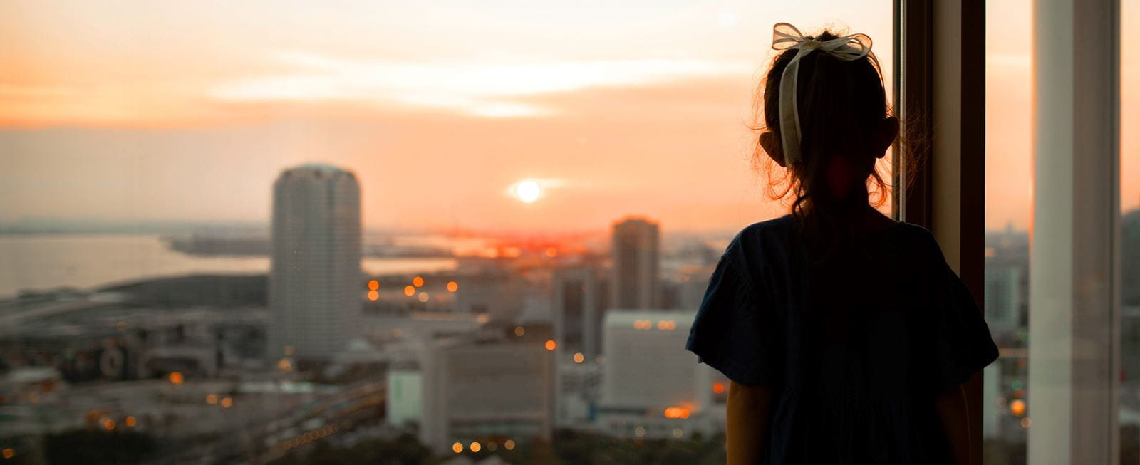 A child looking out of a window at a city skyline during sunset
