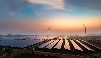 Expansive solar panel field and two wind turbines under a colorful sunrise sky, symbolizing clean energy and sustainability in a serene landscape.